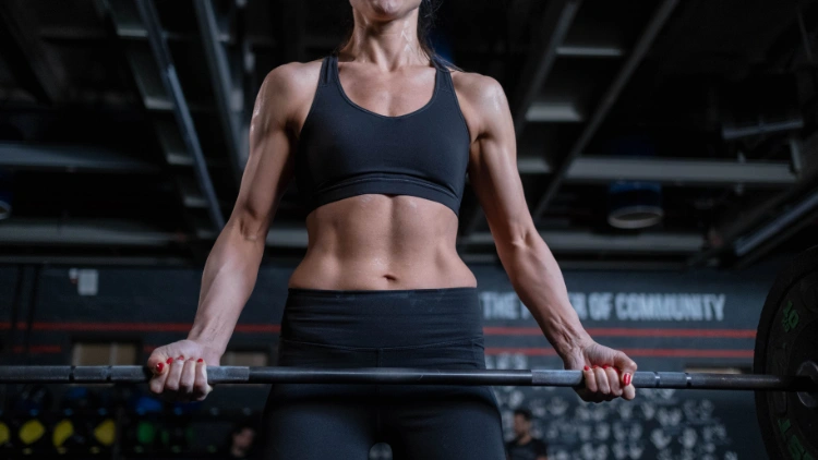 A woman wearing a black tank top and pants showing her core abdominal muscles while lifting a barbell in a gym with black interior.