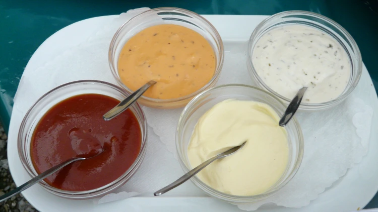 Four bowls of various dressings or dipping sauces, including ketchup, curry sauce, mayonnaise, and ranch dressing, served on a large white plate and placed on a table with a green tablecloth.