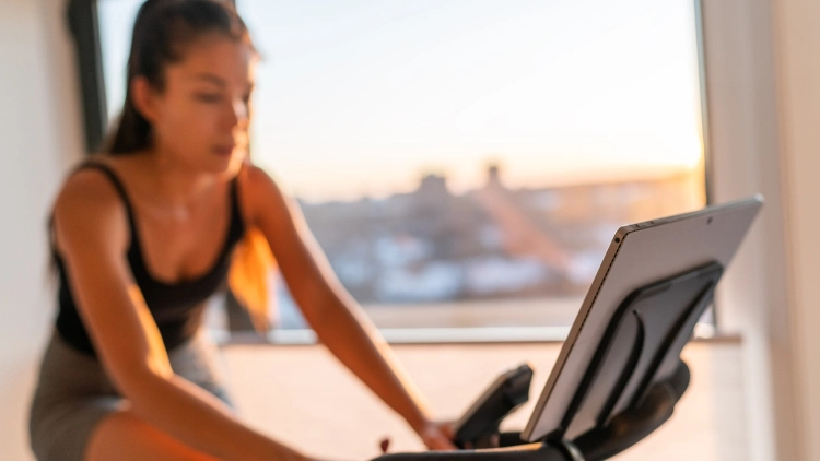 A woman wearing a black tank top and grey short is riding a stationary bike while watching a fitness class at home with a window in the background.