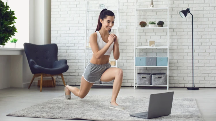 A lady wearing a white tank top, grey shorts, and shoes is doing a workout while looking at a laptop on a grey rug in a room with a plant and a variety of furniture in the background.