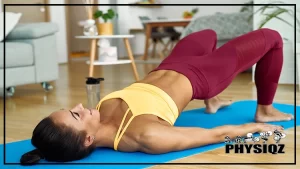 A woman wondering about the glute bridge muscles worked while wearing yellow tank top and red pants as she lifts her hips to perform a glute bridge exercises on a blue yoga mat and inside a living room with wooden floors and various furniture in the background such as a sofa, a side table with flower vase on top, a plant, and a dining table.