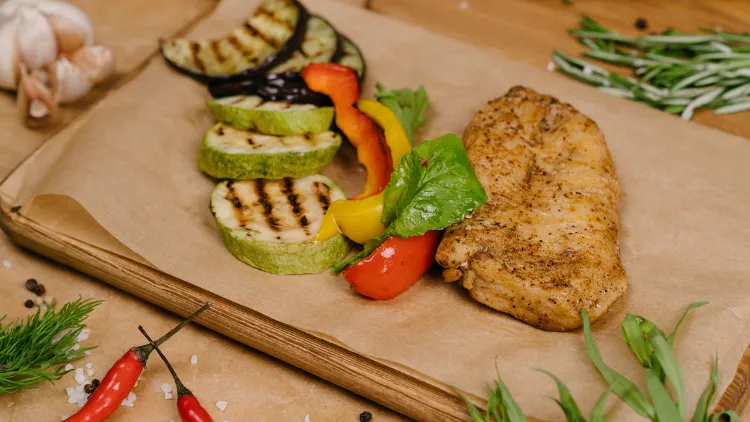 Grilled vegetables and a chicken breast served on a wooden board with table napkin and surrounded by various vegetables such as chili and garlic in the background.