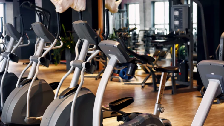 A number of elliptical trainers in a gym with wooden floor and various other machine in the background.