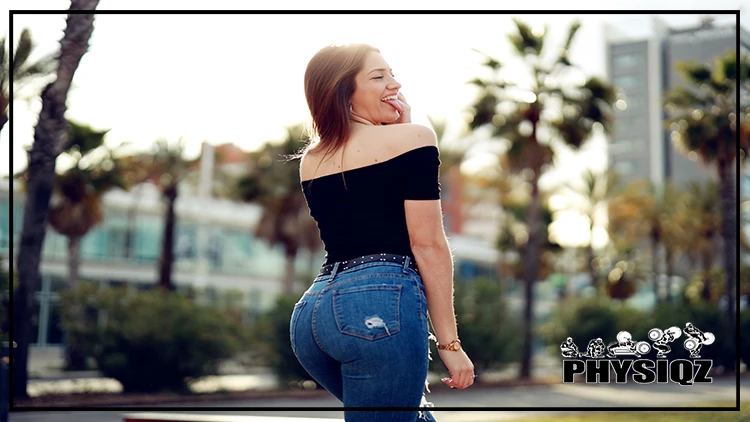 A woman who learned how to get a shelf on the upper glutes is posing with her tongue sticking out, dressed in a black off-shoulder top, denim pants, and a copper colored watch, standing in a bright city landscape that highlights her back side, and there's tall buildings and palm trees in the background.