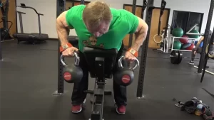 A blonde man wearing a green t-shirt, black pants and red shoes is performing a humble row exercise using a pair of kettlebells in a gym with a treadmill and an exercise ball in the background.