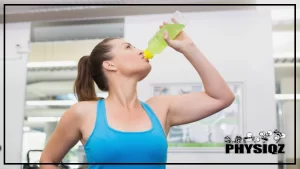 A woman with a ponytail wearing a blue top, holding a plastic bottle of green Mio energy drink while taking a sip.