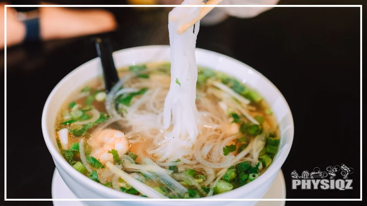 Using chopsticks, a person is grabbing the noodles from a white bowl that has green onions, cilantro, and shrimp in it while wondering is pho keto friendly, or do the noodles and sugary broth have a potential to ruin ketosis?