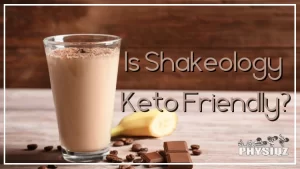 A clear glass filled with a rich, brown chocolate protein shake is shown placed on a wooden table, beside the glass are a sliced banana and a chocolate bar, as well as a small pile of coffee beans, the shake has a thick and creamy texture and is topped with a shake powder, the wooden table has a rustic appearance with visible wood grain, the background is blurred, suggesting that the image was taken in a cozy cafe or kitchen.