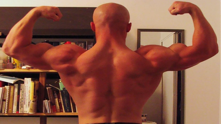 With a book in front of him, a shirtless bald man flexes his muscles on his back and arms. 