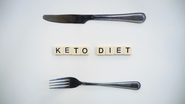 A silver bread knife, a fork and scrabble letter tiles forming the word "keto diet" on top of a white surface.