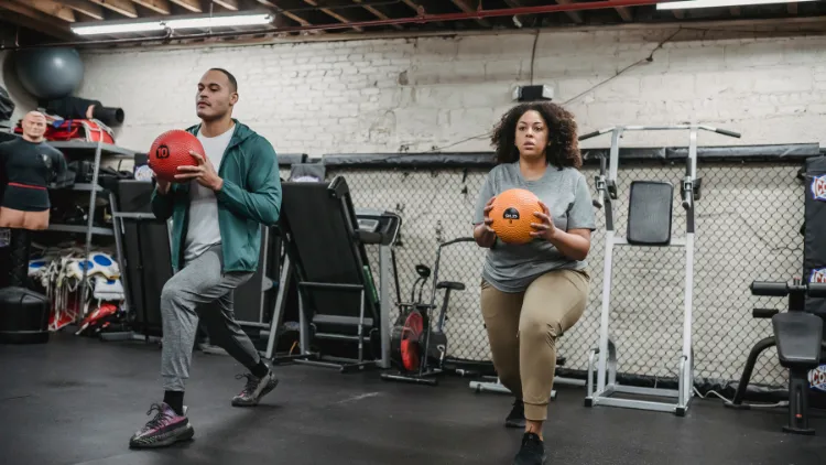 A man wearing green jacket with white shirt under, grey sweatpants, black shoes and a woman wearing grey shirt, khaki pants, and black shoes doing lunges exercise with weight balls in gym with various equipment in the background.