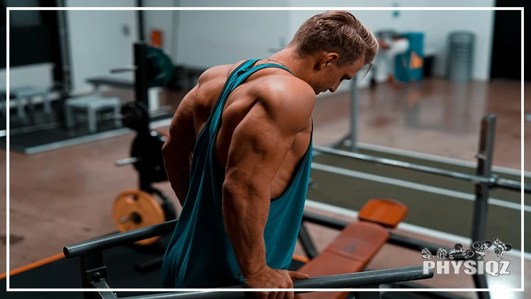 Muscular guy with blonde hair is wearing a blue tank top and getting ready to do dips inside a gym. 
