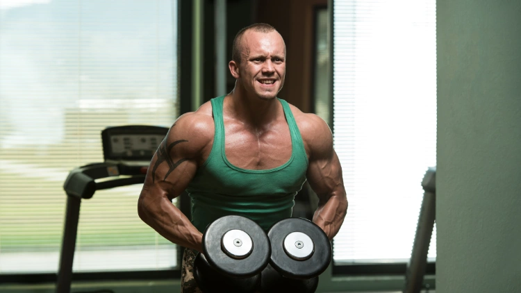 A man with a tattoo on his right arm, wearing a green tank top is performing a dumbbell lateral raise exercise in a gym with a treadmill in the background.