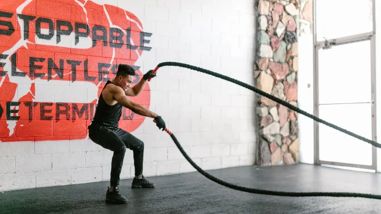 A man in black tank top, pants and shoes with black gym gloves performing an exercise using a black battle rope with red handles in a well-lit studio, with a white wall with an illustration painted on it, stone patterned column, and large window in the background.