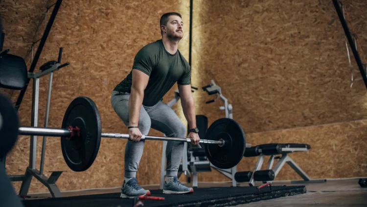 An athletic man in a green shirt and gray pants lifts a barbell loaded with weights, as he stands on a gym floor, behind him, various gym equipment and weight machines can be seen, while he wears a black watch on his wrist.