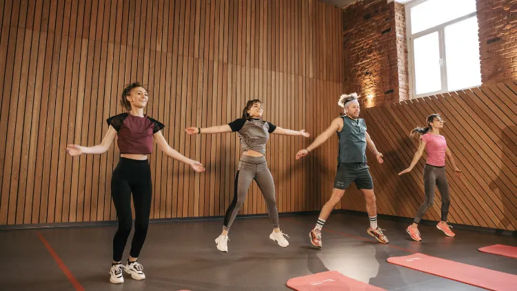 Group of four people doing jumping jacks in a fitness center with wooden slat walling and large window in the background.