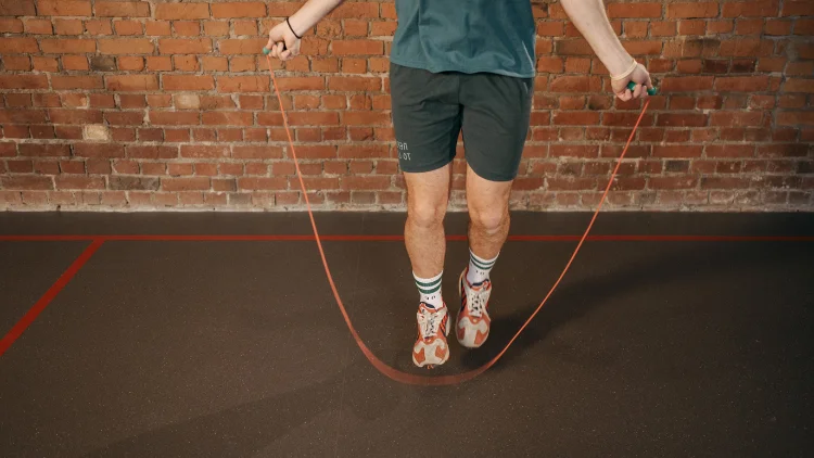 A person, wearing a blue top and black shorts, with orange and white shoes and white socks, is doing an exercise using an orange jump rope in a studio with brick walls in the background.