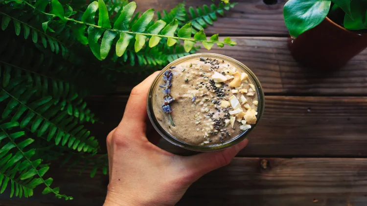 A person holding a glass of smoothie with variety of toppings on it at a table with plants.