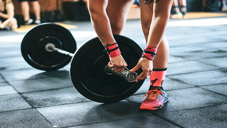 A person, wearing pink wristbands, shoes with matching pink socks, locks a weight plate onto a barbell before doing deadlifts and inside a gym filled with other people in the background.
