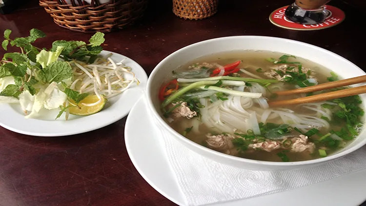 A bowl of soup with vegetables and meat, and some side dish on a plate beside it, served on the table.