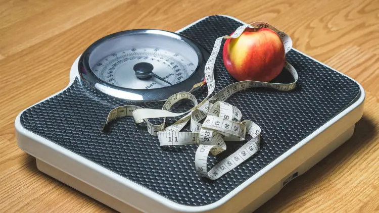 A tape measure and an apple on top of a weighing scale on a wooden surface.