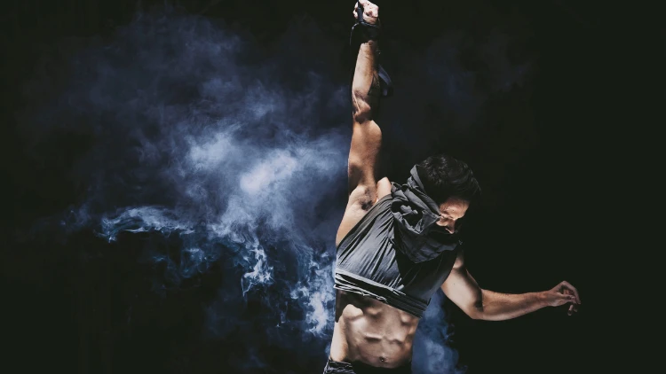 A man with a half-covered face wearing a grey top and black pants is holding on to the rope with smoke in the black background.