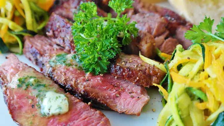 Five slices of steak on top of a white plate with cilantro, zucchini, and cheese on the side. 