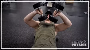 A guy in a green shirt is lying on a black bench inside a gym with black speckled floors and is performing a dumbbell Tate press.