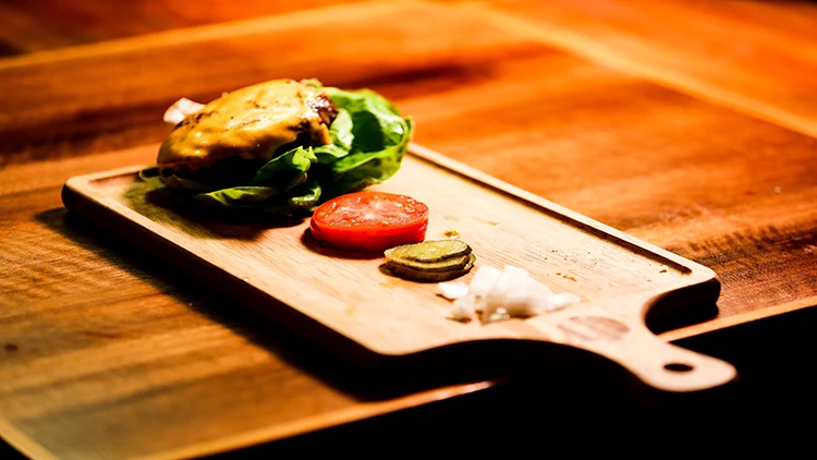A vegan burger, consisting of leafy vegetables, slices of tomato, and other ingredients, is arranged on a wooden board on top of a wooden table.