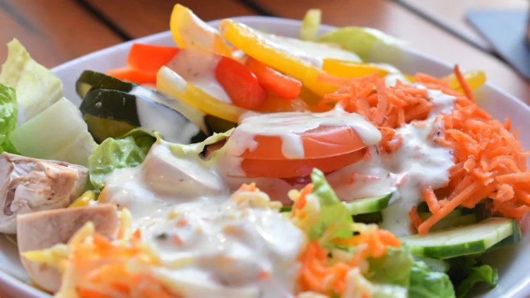 A bowl of vegetable salad that includes ingredients such as shredded carrots, slices of tomatoes, and cucumbers, covered in white dressing, displayed on a wooden table.