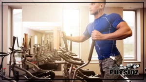 A man with a muscular build, wearing a blue shirt and grey pants can be seen on an elliptical machine inside a gym as he wonders what muscles does the elliptical work, with earphones on and a large window and several other elliptical trainers visible in the background.