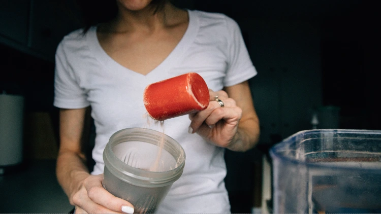 A woman in white crew neck shirt adding a powder to a grey tumbler using a red plastic cup in a dimly lighted room.