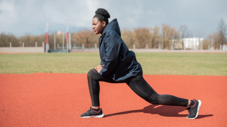 A woman wearing navy blue windbreaker jacket, black pants and black shoes with black socks doing lunge exercise on a red running track with a field in the background.