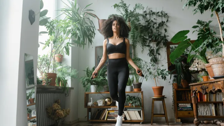 A woman in a matching black tank top and pants, and white shoes, exercises with a jumping rope in a room filled with plants and furniture in the background.