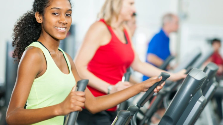 A smiling woman wearing a light green tank top doing an exercise using an elliptical trainer in a gym along with a blonde woman wearing a red tank top and black pants, and a man wearing blue shirt visible in the background.