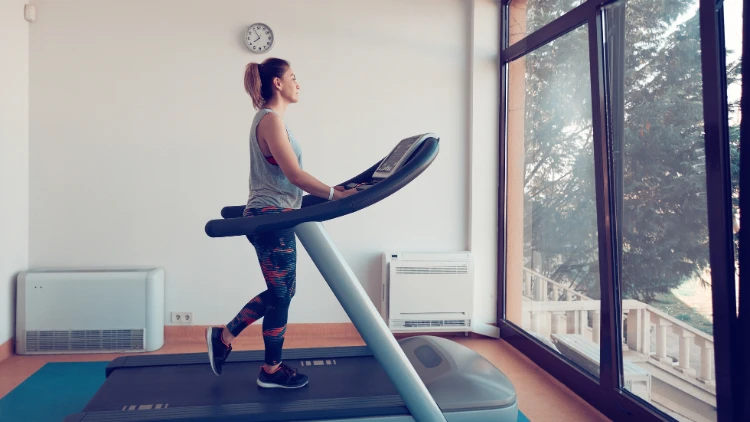 A woman wearing a grey tank top and patterned black leggings, and shoes is walking on a treadmill at home with appliances on the floor such as heater and a wall clock in the background, she is facing towards their glass window with a view of the outdoors.