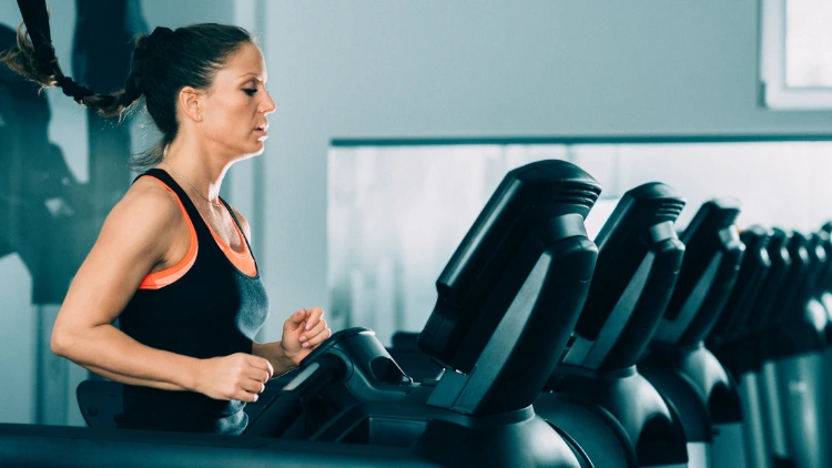 A woman with braided hair, wearing a black and orange tank top is running on a treadmill at a gym with several other treadmills in the background.