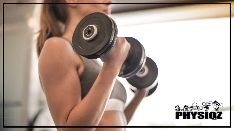 While following the 12 week dumbbell workout plan PDF, a white woman with a blonde pony tail is in a grey sleeveless top that has a white band on the bottom and performing dumbbell curls and the background is blurred out.