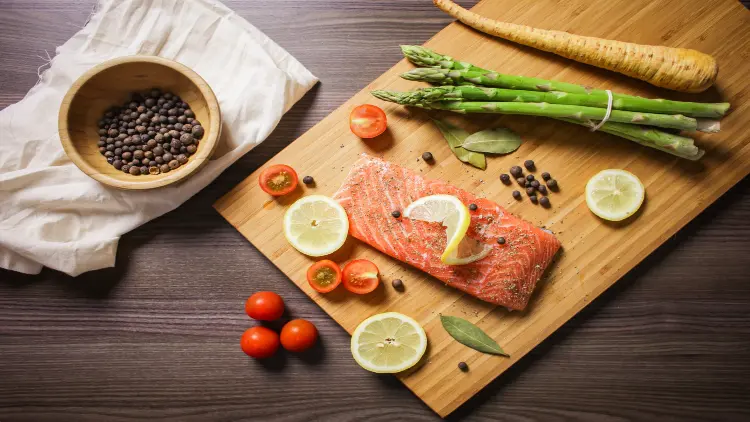 A piece of turnip, a bundle of asparagus, slices of lemon and tomatoes, a slice of raw salmon with black pepper set on a wooden board, and to the side of this arrangement is a bowl of black pepper placed on top of a white fabric, all of which are presented on a surface made of dark wood.