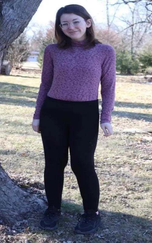 A lady wearing eyeglasses, purple long sleeves, black tights, and black shoes is standing beside a tree, showing her body, which is slimmer and more defined, with green grass and trees in the background.