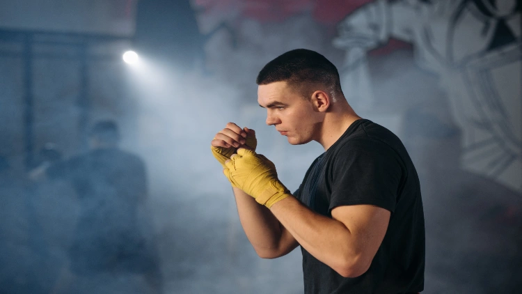 A man in a black shirt and yellow boxing bandages wrapped over both hands is shown shadowboxing in a smoke-filled gym with another man boxing in the background.