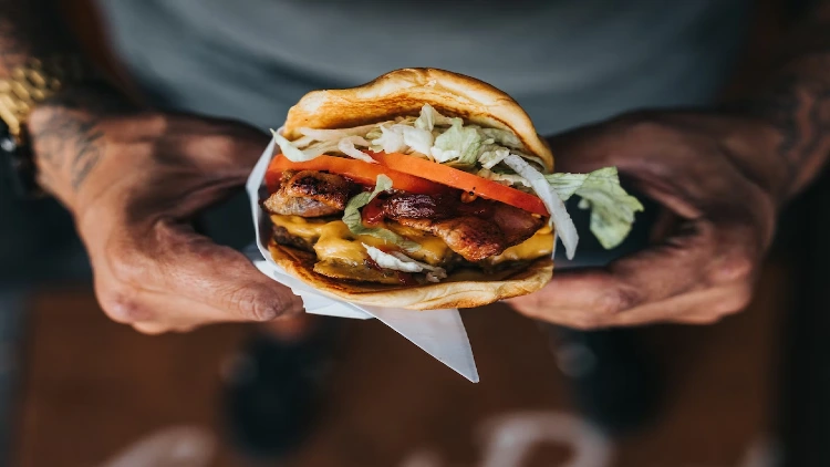 A man with both hands grasping a delicious burger that is loaded with fresh tomato and lettuce, juicy meat, and melted cheese, the burger is neatly arranged and looks mouthwateringly delicious.
