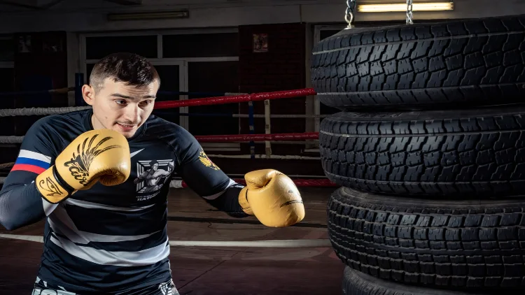 A man wearing black, white, blue, and red long sleeves and yellow boxing gloves is boxing in three-layered black car tires in a gym with a boxing ring visible in the background.