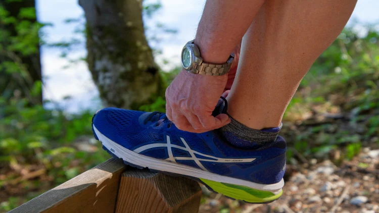 A man's hand wearing a watch is tying the laces of his blue shoe on a wooden surface, with a forest-like background in the background.