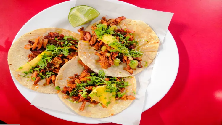 Three tacos filled with ground beef, beans, lettuce, lemon, and cheese slices on a white table napkin and plate displayed on a shiny red surface.