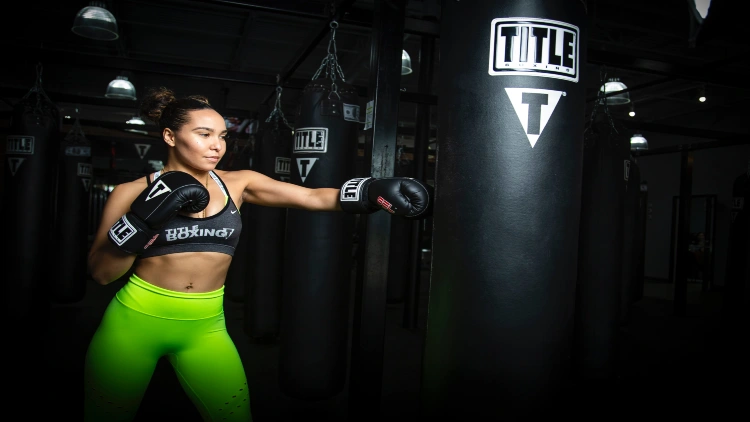 A woman with her hair styled in a bun is seen wearing a gray tank top and neon green tights, while wearing black boxing gloves and engaging in boxing with a black punching bag, in a dimly lit gym with several punching bags in the background.