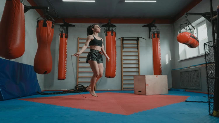 A girl with ponytail wearing a black gym tank top and black and white shorts is shown doing jumping ropes in a gym with a blue and red floor, multiple red punching bags hanging in the background, and a wooden plyo bench.
