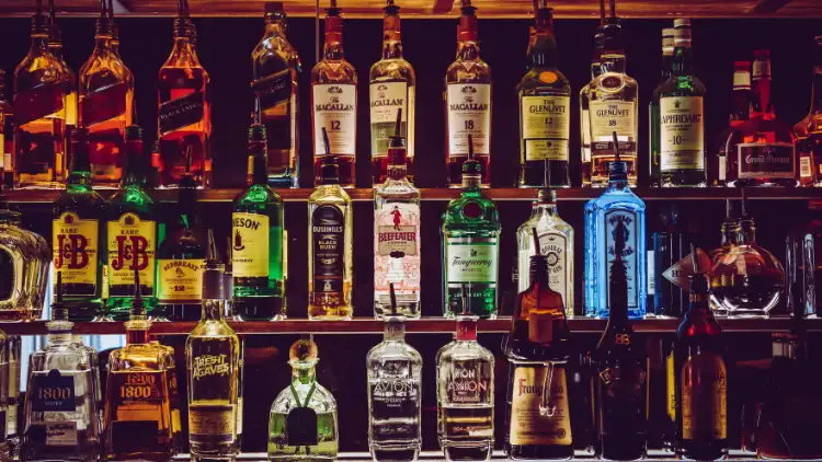 A bar has a shelf full of various alcoholic drinks, ranging from whiskey to gin, rum, tequila, and vodka, arranged in neat rows with some bottles prominently displayed in the front, creating a simple but vibrant and colorful focal point.