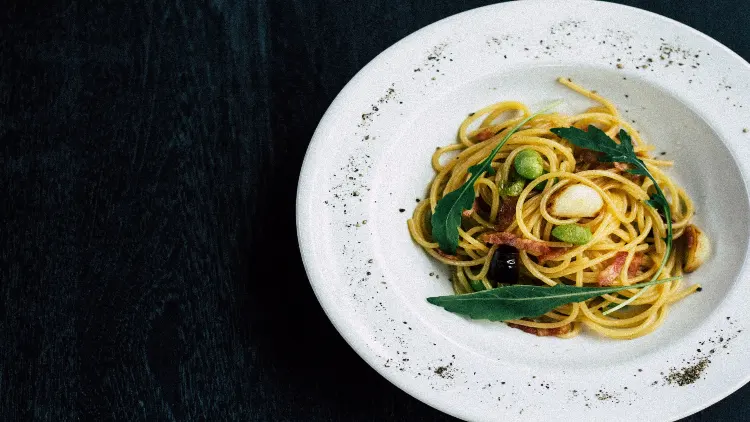 Flavorful spaghetti noodles seasoned with ground pepper, along with crispy bacon, mushrooms, and fresh basil, served on a white plate placed on a black table-like surface.