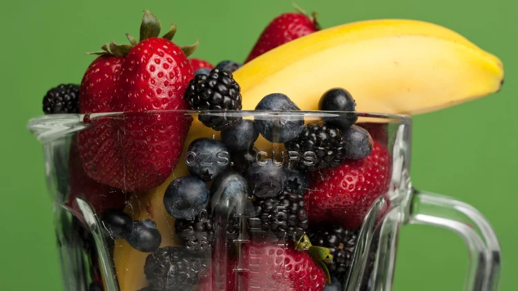 A blender filled with a vibrant mix of fresh fruits, including bananas, blueberries, and black raspberries, the fruits are bright and colorful, with the blues and purples of the blueberries and black raspberries contrasting against the yellow of the bananas.